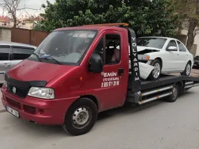 Ağaçlı bir sokakta ön kısmı ezilmiş beyaz sedanı kasasında taşıyan Emin Oto Kurtarma çekicisi