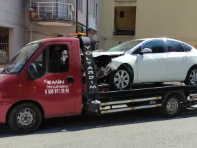 Apartman önünde ön tarafı kazada hasar görmüş beyaz sedanı taşıyan Emin Oto Kurtarma çekicisi