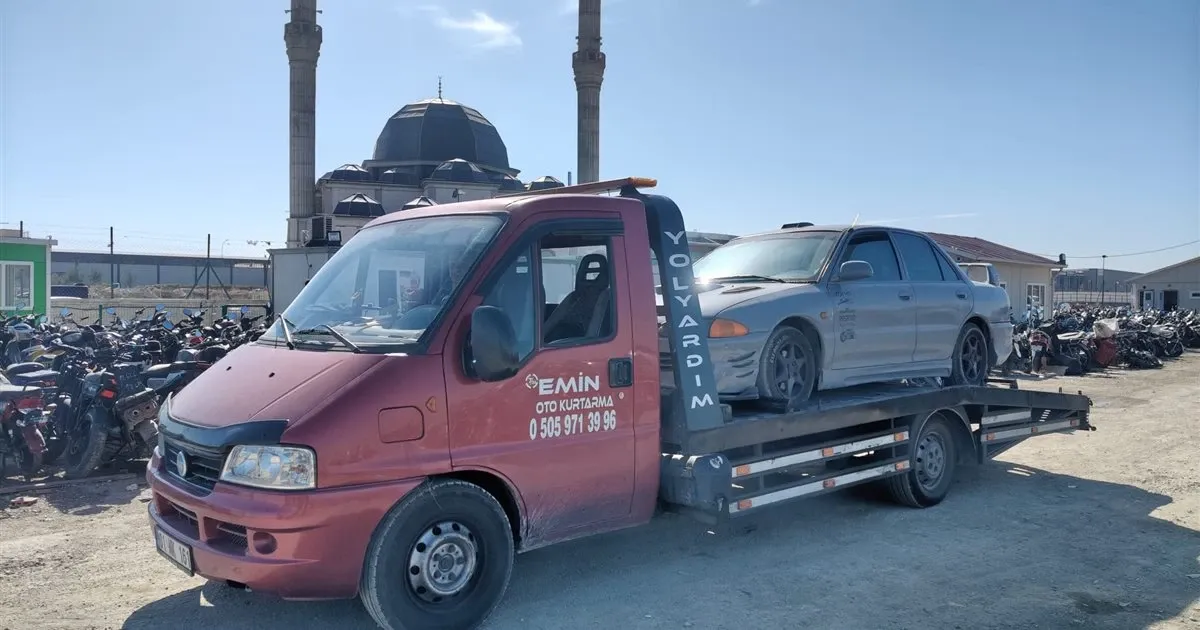 Arkasında iki minareli cami bulunan açık otoparkta gri sedan yüklü kırmızı Emin Oto Kurtarma çekicisi