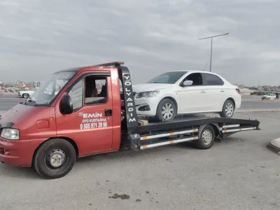 Bulutlu havada geniş cadde kenarında beyaz sedanı taşıyan kırmızı Emin Oto Kurtarma çekicisi
