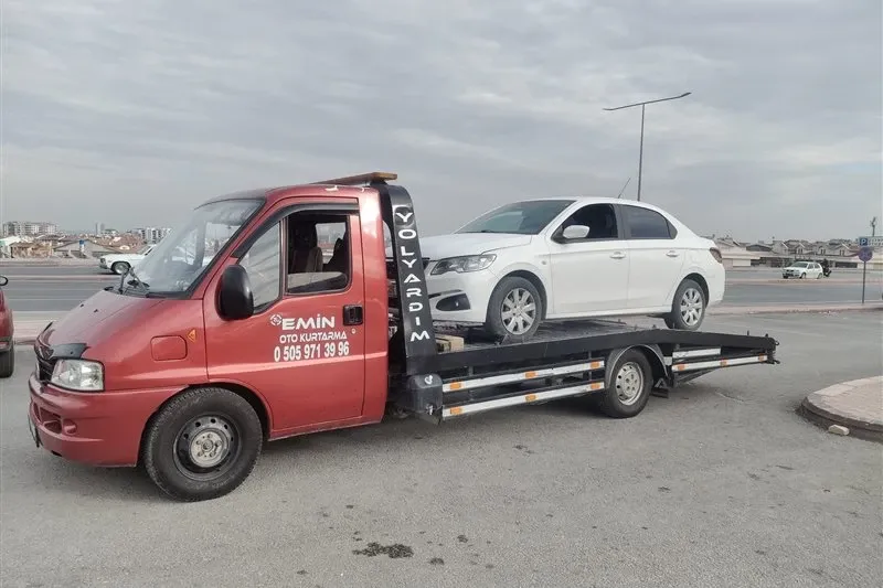 Bulutlu havada geniş cadde kenarında beyaz sedanı taşıyan kırmızı Emin Oto Kurtarma çekicisi