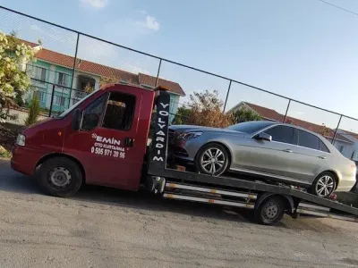Güneşli bir günde çiçekli bahçe ve tel örgü yanında gümüş renkli sedanı taşıyan Emin Oto Kurtarma çekicisi