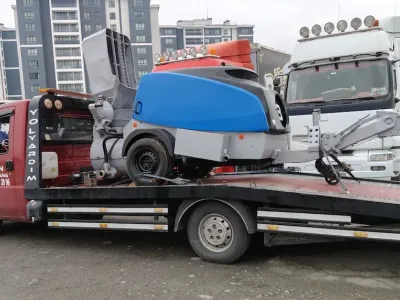 Yüksek apartmanlar arasında mavi gövdeli tekerlekli inşaat makinesini taşıyan Emin Oto Kurtarma çekicisi